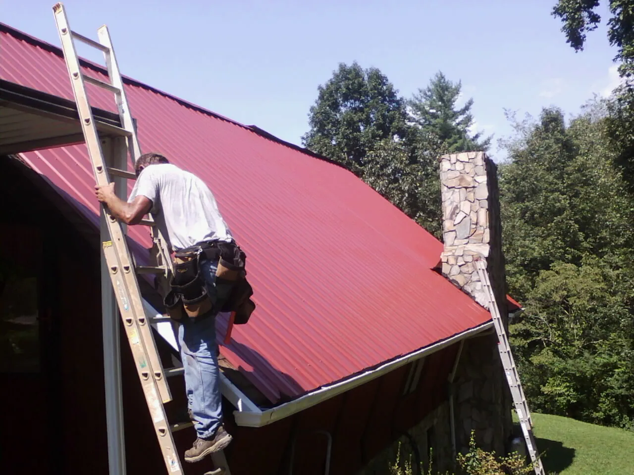 Roofer on a ladder beside a red metal roof and stone chimney — Soffit & Fascia Repair by Roofing Brothers in Sanger